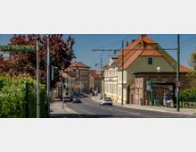 Straßenszene mit Wohn- und Geschäftshäusern im historischen Stil, teils mit gelb gestrichenen Fassaden und roten Ziegeldächern. Im Vordergrund verlaufen Gehwege, Fahrbahnen, Verkehrszeichen und Straßenbeleuchtung, umgeben von einzelnen Bäumen und einem Bushaltestellenschild.