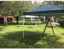 Grüne Wiese mit aufgebauten Pavillons und Tischen, die mit weißen Tischdecken bedeckt sind. Im Hintergrund sind ein rotes Gebäude, Bäume und Büsche zu sehen, und eine Holzleiter steht aufrecht im Vordergrund.