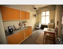 Schlichte, leicht abgenutzte Küche mit orangefarbenen Schrankfronten, langer Arbeitsplatte und frei stehendem Herd neben einem großen Fenster. Im Raum stehen ein kleiner Esstisch mit Stühlen, ein alter Kühlschrank und einfache Küchenutensilien an der Wand.