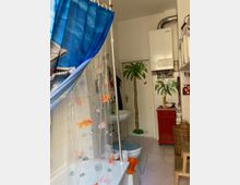 Badezimmer mit Badewanne und goldfischbedrucktem Duschvorhang. Im Raum sind eine rote Kommode, ein an der Wand montierter Gasboiler, ein geflochtener Wäschekorb und tropische Dekorationen wie gemalte Palmen an der Tür sichtbar.