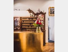 Eleganter Innenraum mit einer goldfarbenen Empfangstheke, dahinter ein antikes Holzregal mit kleinen Fächern und dekorativen Gegenständen. Auf der Theke steht eine Vase mit roten und weißen Blumen.