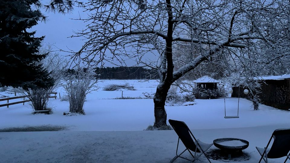 Verschneiter Garten mit großem, schneebedecktem Baum im Vordergrund und Blick auf ein weißes Feld und einen dunklen Waldrand in der Ferne. Rechts stehen zwei Stühle und ein niedriger Tisch im Schnee, daneben eine Schaukel und ein kleines Gartenhäuschen.