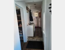 Schmaler, schlichter Flur in einem Wohnhaus mit weiß gestrichenen Wänden, einer Garderobe an der linken Wand und einem kleinen weißen Regal. Der Boden ist mit Fliesen versehen, und im Hintergrund führt eine Tür mit Milchglas und einem Stoppschild-Aufkleber zu einem weiteren Raum. Links befindet sich ein Schuhregal in einer Nische.