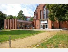 Backsteingebäude mit gotischen Fenstern, ergänzt durch einen modernen Anbau mit Glasfront und rotem Klinker. Vor dem Gebäude eine Grünfläche mit Kiesweg und außen aufgestellte Sitzmöbel.