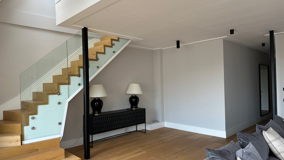 Modernes Wohninterieur mit offener Holztreppe und Glasgeländer, die zu einer Galerieebene führt. Heller Raum mit Holzfußboden, weiß-grauen Wänden, schmalen Deckenleuchten und einer schwarzen Konsole mit zwei Tischlampen.