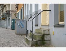 Straßenszene mit einer grauen Gebäudefassade, einer kurzen Treppe mit Metallgeländer und einem Gehweg aus Kopfsteinpflaster. Im Hintergrund sind Balkone, ein Schild mit der Aufschrift „Café“ und eine Laterne sichtbar, während eine Mauer mit Graffiti den Vordergrund prägt.