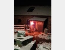 Schneebedeckter Hof vor einem Backsteingebäude mit geneigtem Dach und Dachfenster; ein Eingang ist beleuchtet, über dem ein rotes Leuchtsymbol in Form eines Sterns hängt. Im Vordergrund sind Holzzäune und ein schneebedecktes Betonelement zu sehen.