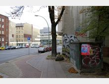 Straßenszene in einer städtischen Umgebung mit mehrstöckigen Gebäuden im Hintergrund; an der rechten Seite des Gehwegs befindet sich ein Baum sowie ein Stromkasten mit Graffiti und Plakaten, während links Autos entlang einer ruhigen Straße parken.