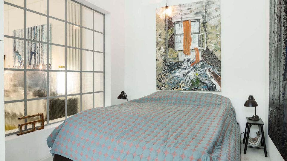 Minimalist bedroom with white walls, a double bed covered in a blue patterned quilt, and black bedside lamps on small tables. A large frosted grid-style window and a colorful abstract painting above the headboard define the space.