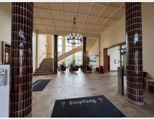 Großer Eingangsbereich mit hohen, türkis-schwarzen Kachelsäulen, einem cremefarbenen Kassettendecken-Design und einem Treppenaufgang links. Zentral steht ein Kronleuchter, und im Hintergrund befinden sich Sitzbereiche mit Pflanzen vor großen Fenstern.