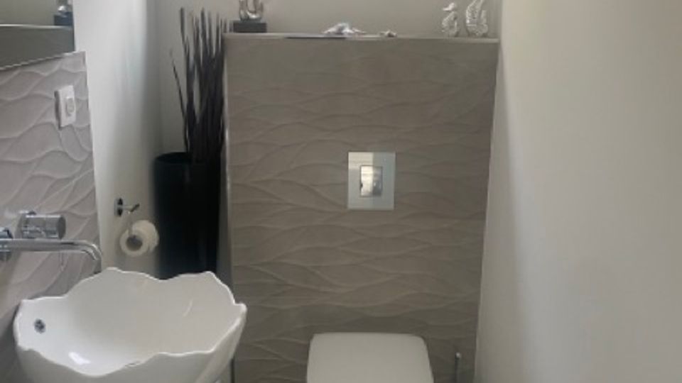 Schmales, modernes Gäste-WC mit wandhängender Toilette, kleiner Waschschale auf Unterschrank und grau strukturierten Wandfliesen. Am Ende des Raums sitzt ein Fenster über der Vorwandinstallation, davor dekorative Elemente und hohe Ziergräser.