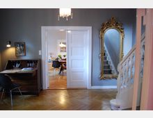 Wohnraum mit Parkettboden, grauen Wänden und einer weißen Tür, die zu einem angrenzenden Esszimmer führt; links ein Holzschreibtisch mit Stuhl, rechts eine geschwungene Treppe mit weißem Geländer und ein großer, goldgerahmter Spiegel.