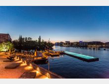 Eine Uferpromenade mit beleuchteten Holzstegen und einem länglichen, schwimmenden Pool auf einem Fluss bei Dämmerung. Im Hintergrund sind moderne Gebäude und beleuchtete Fenster zu sehen, sowie Bäume und ein Kran.