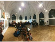 Ein heller Raum mit gotischen Spitzbögen und großen Fenstern, in dem eine Veranstaltung stattfindet. An der linken Wand hängen Gemälde, im Vordergrund ein Flügel, umgeben von sitzendem Publikum auf Holz- und Stuhlreihen.