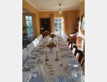Langer Speisesaal mit großem, weiß gedecktem Banketttisch, zahlreichen Gedecken, Kristallgläsern mit Goldrand, Kerzenleuchtern und Blumenarrangements. Helle Wände, hohe Fenster und eine Glastür mit Vorhängen sowie ein Kamin und ein großes gerahmtes Porträt prägen den Raum.
