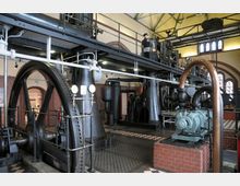 Innenraum einer historischen Maschinenhalle mit großen Zahnrädern, Metallrohren, Manometern und Dampfmaschinen. Der Raum hat Ziegelwände, ein Muster aus gefliesten Böden und hohe Fenster mit Rundbögen.