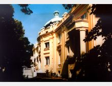 Historisches Gebäude mit neoklassizistischen Elementen, gekennzeichnet durch eine beige Fassade mit Säulen und reich verzierten Fenstern, sowie einer Kuppel im Hintergrund; das Gebäude ist von Bäumen umgeben.