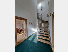 Ein Eingangsbereich mit einer modernen, offenen Holztreppe und Metallgeländer, die nach oben führt. Links befindet sich eine offene Tür mit Holzrahmen, die zu einem Raum mit Esstisch und Stühlen führt, während der Bereich mit weiß gestrichenen Wänden und einem dunkelgrünen Boden gestaltet ist.