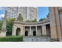 Steinernes, halbkreisförmiges Bauwerk mit Säulen und Bögen, verziert mit Reliefs und Skulpturen, im Vordergrund ein Obelisk, dahinter Grünflächen und ein modernes, hohes Wohngebäude.