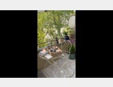 Ein kleiner Balkon mit verschnörkeltem weißen Metalltisch und Stühlen, darauf eine Kerze, Bücher und ein Tablett mit Getränken. Im Hintergrund grüne Pflanzen und Bäume, dekoriert mit Blumentöpfen und einer Milchglas-Abtrennung.