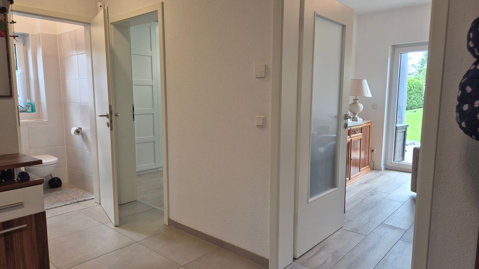 Heller Flur in einer Wohnung mit grauen Bodenfliesen und weißen Wänden; links führt eine offene Tür in ein weiß gefliestes Badezimmer. Rechts öffnet sich der Flur zu einem Wohnraum mit Glastür, Holzmöbel und einer großen Terrassentür mit Blick ins Grüne.