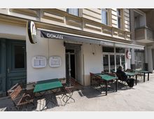 Straßencafé vor einem Restaurant in einem Altbau mit dekorierter Fassade, Markise mit der Aufschrift „GOKAN“ und großen Fenstern. Davor stehen mehrere grüne Tische mit Holzstühlen auf einem gepflasterten Gehweg.