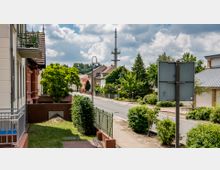 Wohnstraße in einer Kleinstadt mit niedrigen Wohnhäusern, Vorgärten und einem Gehweg, gesäumt von Bäumen und Grünflächen. Im Hintergrund ein hoher Funkmast und dichter Baumbestand unter leicht bewölktem Himmel.