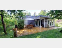 Einstöckiges Haus mit grauer Holzfassade und blauem Ziegeldach, umgeben von Bäumen und Garten. Im Vordergrund eine Terrasse aus gelben Steinplatten mit Gartenmöbeln und Pflanzen.