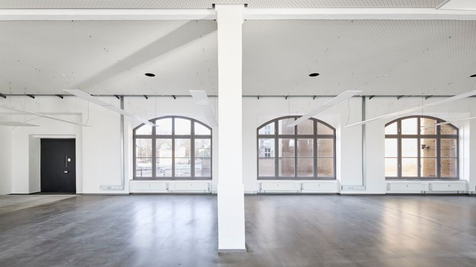 Großer, leerer Innenraum mit einem modernen, grauen Boden, weißen Wänden und einer Lochdecke mit eingelassenen Leuchten. Große Bogenfenster lassen viel Tageslicht in den Raum, und eine weiße Säule steht mittig im Vordergrund.