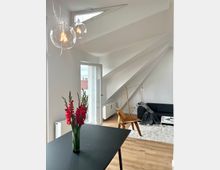 Helles Wohnzimmer in einer Dachgeschosswohnung mit schrägen weißen Wänden und Holzboden. Zu sehen sind ein schwarzer Tisch mit roten Blumen, ein Holzstuhl, ein schwarzes Sofa auf einem weißen Teppich, moderne Hängeleuchten und eine offene Balkontür mit Blick auf Dächer.