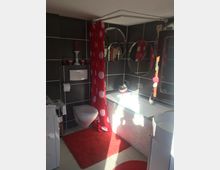 Badezimmer mit dunklen Fliesenwänden, einer weißen Toilette an der Wandmontage, einer Badewanne mit Mosaikverkleidung und einem roten Duschvorhang mit weißen Punkten. Rote Akzente wie ein Teppich und Kerzen ergänzen das Design, daneben befinden sich runde Wandspiegel und eine Waschmaschine.