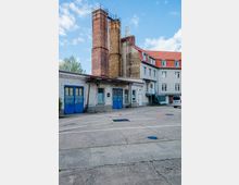 Hinterhof eines Gebäudekomplexes mit grauen Fassaden, blauen Garagentoren und drei hohen, teils verwitterten Ziegelschornsteinen. Im Hintergrund ist ein mehrstöckiges Wohn- oder Bürogebäude mit roten Dächern und Dachfenstern zu sehen.