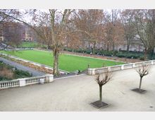 Weitläufige Parkanlage mit einer zentralen rechteckigen Rasenfläche, umgeben von Gehwegen und niedrigen weißen Balustraden. Im Vordergrund stehen mehrere beschnittene Bäume, während große, kahle Bäume den Hintergrund dominieren.