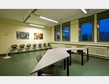 Ein schlichter Besprechungsraum mit grünem Boden, gelblichen Wänden und drei großen Fenstern. An der Wand befinden sich zwei Bilder, darunter eine Reihe von Stühlen, und in der Mitte steht ein dreieckiger Konferenztisch mit weiteren Stühlen.