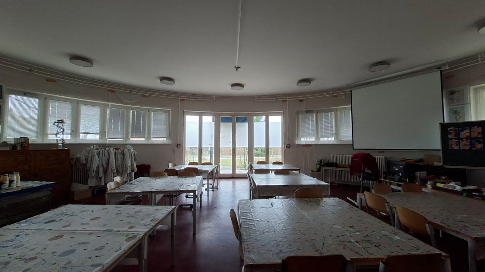 Heller Kunstraum oder Klassenraum mit mehreren großen Arbeitstischen und Stühlen, einer gebogenen Fensterfront mit Jalousien und einer zentralen Glastür nach draußen. Rechts hängt eine große Projektionsleinwand; an der Wand stehen Regale und Materialien, die Decke ist mit runden Leuchten ausgestattet.