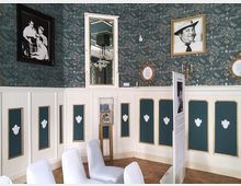 Ein eleganter Innenraum mit blumengemusterter Wandtapete in Dunkelgrün, weißer Holzvertäfelung mit goldenen Details und weißen Zierreliefs. An den Wänden hängen gerahmte Fotografien und Spiegel, und im Raum stehen mit weißen Hussen bezogene Stühle sowie eine Infotafel.