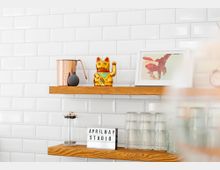 Zwei Holzregale vor einer weißen Wand mit rechteckigen, glänzenden Fliesen. Auf den Regalen stehen ein goldener Winkekater, ein Kupferkrug, ein gerahmtes Bild, ein Pressfilter, gestapelte Gläser und ein Lichtkasten mit der Aufschrift "APRIL MAY STUDIO".