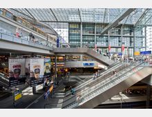 Innenansicht eines modernen Bahnhofes mit mehreren Ebenen und gläserner Dachkonstruktion. Rolltreppen und Fußgängerbrücken verbinden die Ebenen, während Geschäfte und Werbeplakate sichtbar sind; im Hintergrund sind Gleise erkennbar.