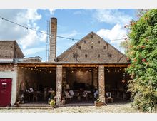 Offener, überdachter Bereich eines alten Backsteingebäudes mit Holzbalkendecke und Säulen, dekoriert mit Lichterketten. Im Hintergrund befinden sich rustikale Möbel und Dekorationen, daneben ist eine Begrünung mit rot blühenden Pflanzen zu sehen.