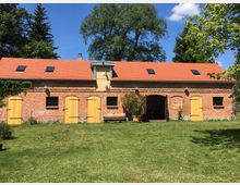 Ziegelsteingebäude mit rotem Satteldach und mehreren gelben Holztüren, eingebettet in eine grüne Rasenfläche; dahinter stehen große Bäume, unter blauem Himmel.