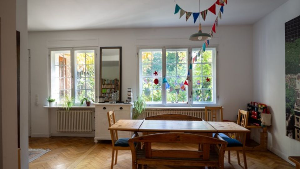 Heller Essbereich mit Holztisch und Stühlen auf Fischgrätparkett, vor großen Sprossenfenstern mit Blick ins Grüne. An der Decke hängen bunte Wimpelgirlanden, daneben stehen eine Kommode mit Spiegel und Zimmerpflanzen.