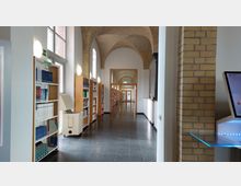 Ein langer Bibliotheksflur mit Regalen voller Bücher auf der linken Seite und einer Backsteinbogendecke. Helle Wandleuchten und große Fenster sorgen für viel Licht, während am Ende des Flurs eine Glastür zu sehen ist.