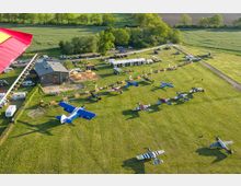 Eine Luftaufnahme eines kleinen Flugplatzes mit einem offenen Feld, auf dem mehrere bunte Leichtflugzeuge geparkt sind. Im Hintergrund stehen ein Gebäude, Zelte und geparkte Fahrzeuge, umgeben von Bäumen und landwirtschaftlichen Flächen.