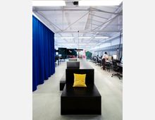 Großraumbüro mit modernen Möbeln und offener Deckenstruktur. Im Vordergrund steht eine schwarze Sitzbank mit gelbem Kissen, im Hintergrund sind Arbeitsplätze mit Computern und blauen sowie grünen Raumtrennern sichtbar.