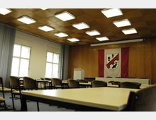 Konferenzraum mit Holzwänden und einer Holzdeckenverkleidung, helle rechteckige Leuchten in der Decke. Vorne ein Rednerpult und eine große Fahne mit Wappen, dahinter mehrere Sitzgelegenheiten; links Fenster mit weißen Gardinen.