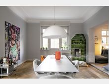 Esszimmer mit modernem Design, einem großen weißen Tisch und grauen Stühlen, einer Hängelampe in Kugelform, einem grünen, gefliesten Kachelofen und einem großen Fenster im Hintergrund. An der Wand hängt ein großformatiges, buntes Kunstwerk, und ein Servierwagen steht seitlich vom Tisch.