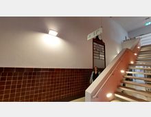 Treppenhaus mit rot-braunen Kacheln an den Wänden, moderner Beleuchtung und einer metallischen Treppe mit Handlauf. An der Wand hängt eine historische Zuganzeigetafel mit einer Uhr, darüber ein Wegweiser.