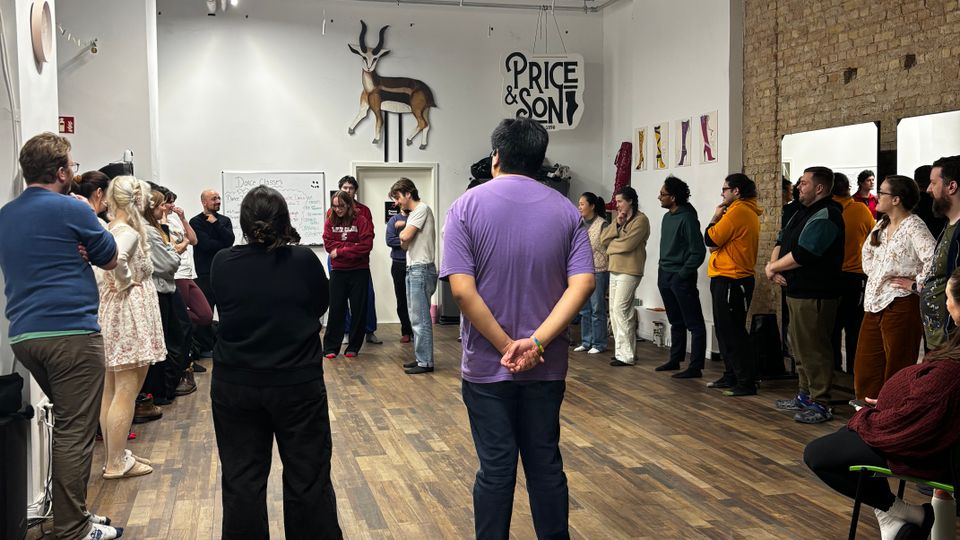 Large open-plan studio or event space with wood plank flooring, white walls, and an exposed brick wall, featuring a grid of ceiling panels with visible pipes and strip lighting. Artwork and signage hang on the walls, including a mounted antelope sculpture above a doorway and a “Price & Son” graphic.