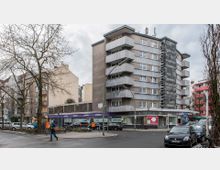 Mehrstöckiges Wohn- und Geschäftsgebäude an einer Straßenecke mit Balkonreihen und Fassaden aus Beton und dunklen Platten. Im Erdgeschoss befinden sich mehrere Ladenlokale mit großen Schaufenstern, auf dem Gehweg sind parkende Fahrräder und Autos sichtbar, sowie einige Passanten.