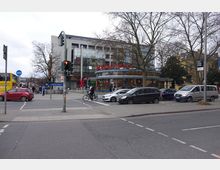 Straßenkreuzung in einer Stadt mit mehreren parkenden und fahrenden Autos, Fußgängern und einem Fahrradfahrer. Im Hintergrund befinden sich ein modernes Gebäude mit Glasfassade, ein Café mit dem Schriftzug „BackFactory“ und ein U-Bahn-Eingang mit der Beschriftung „Rathaus Steglitz“.